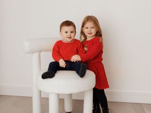 Two young children in matching red outfits pose on a white chair for a holiday portrait.