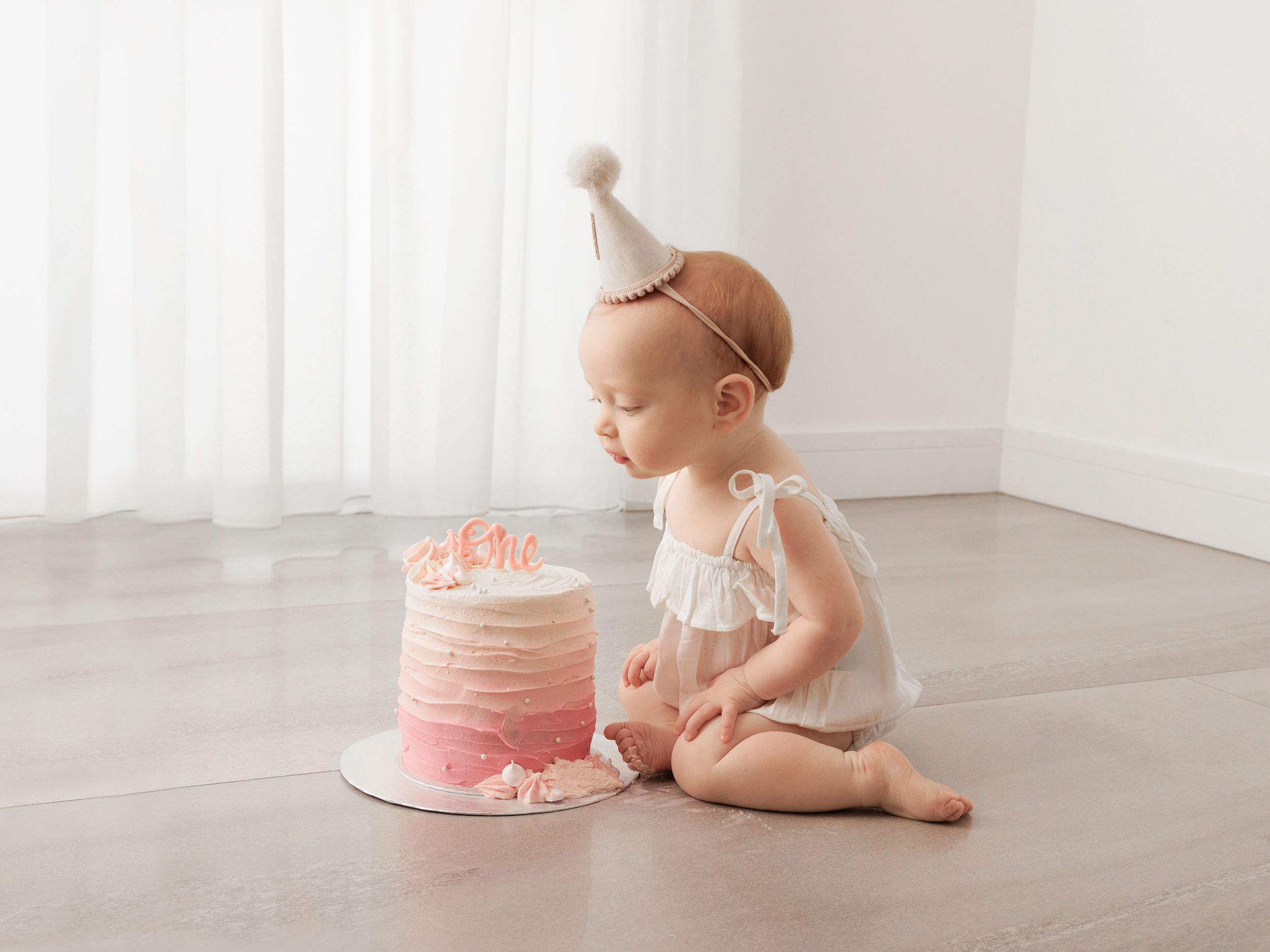 Baby girl in white outfit and party hat sits on floor looking at pink ombre first birthday smash cake.