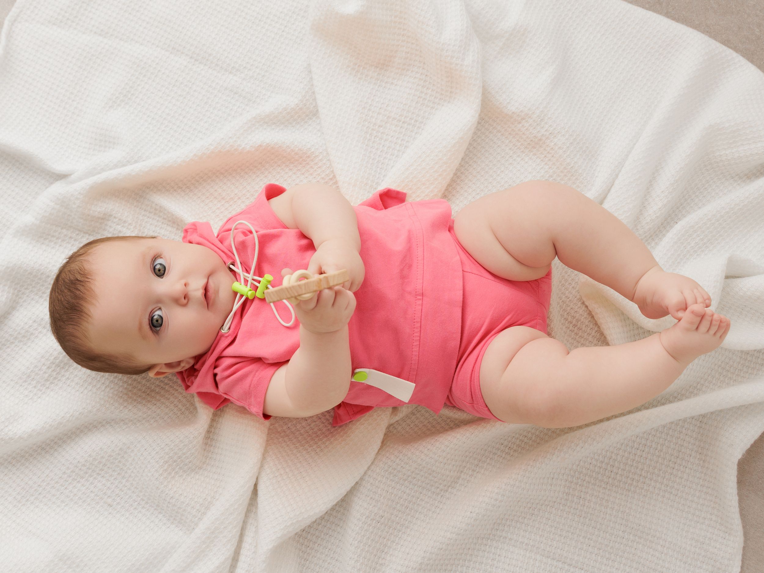 Adorable baby girl in pink onesie lying on white blanket, holding a colorful toy while looking up curiously.