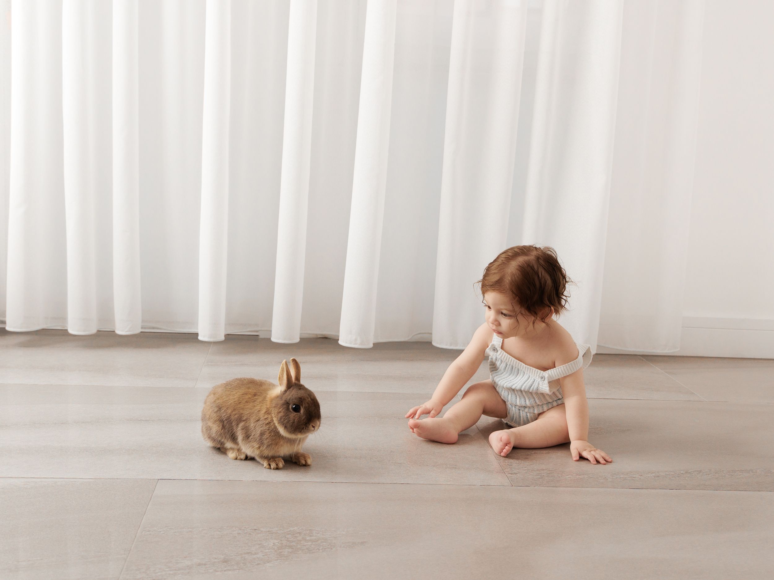 A baby girl in a floral romper sits on a light floor, curiously watching a small brown bunny rabbit indoors.