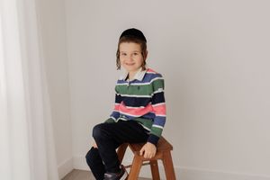 A child in a striped multicolor sweater sits on a wooden stool against a white wall.
