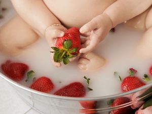 Baby sitting in a metal tub filled with milk and fresh strawberries, holding a strawberry with tiny hands.