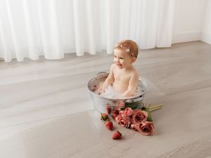 Baby girl sits in a small metal tub with bubbles, roses, and strawberries on a light wood floor.