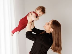A young mother joyfully lifts her smiling baby in a red onesie above her head in a bright white room.