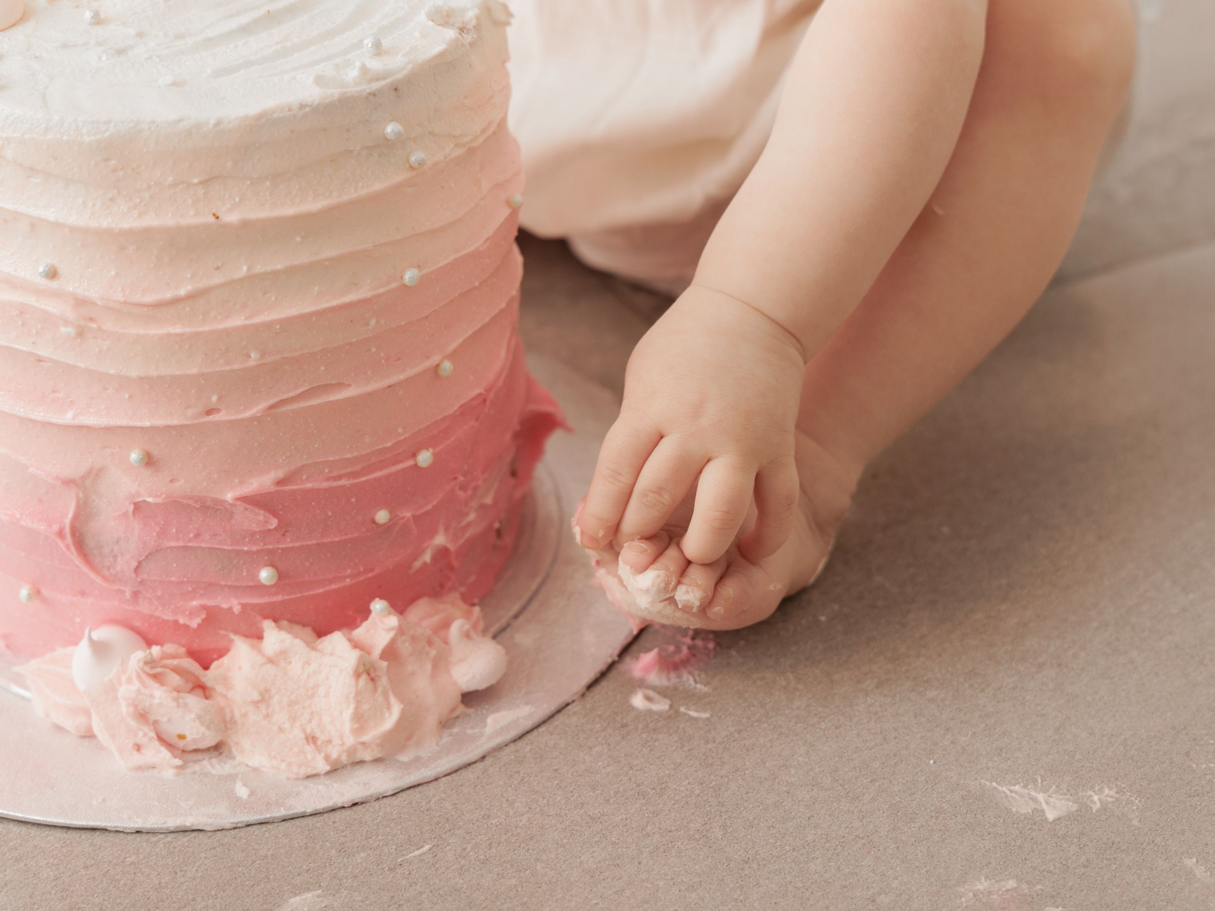 Baby hands touching a pink ombre smash cake with white frosting and pearl decorations on a gray background.