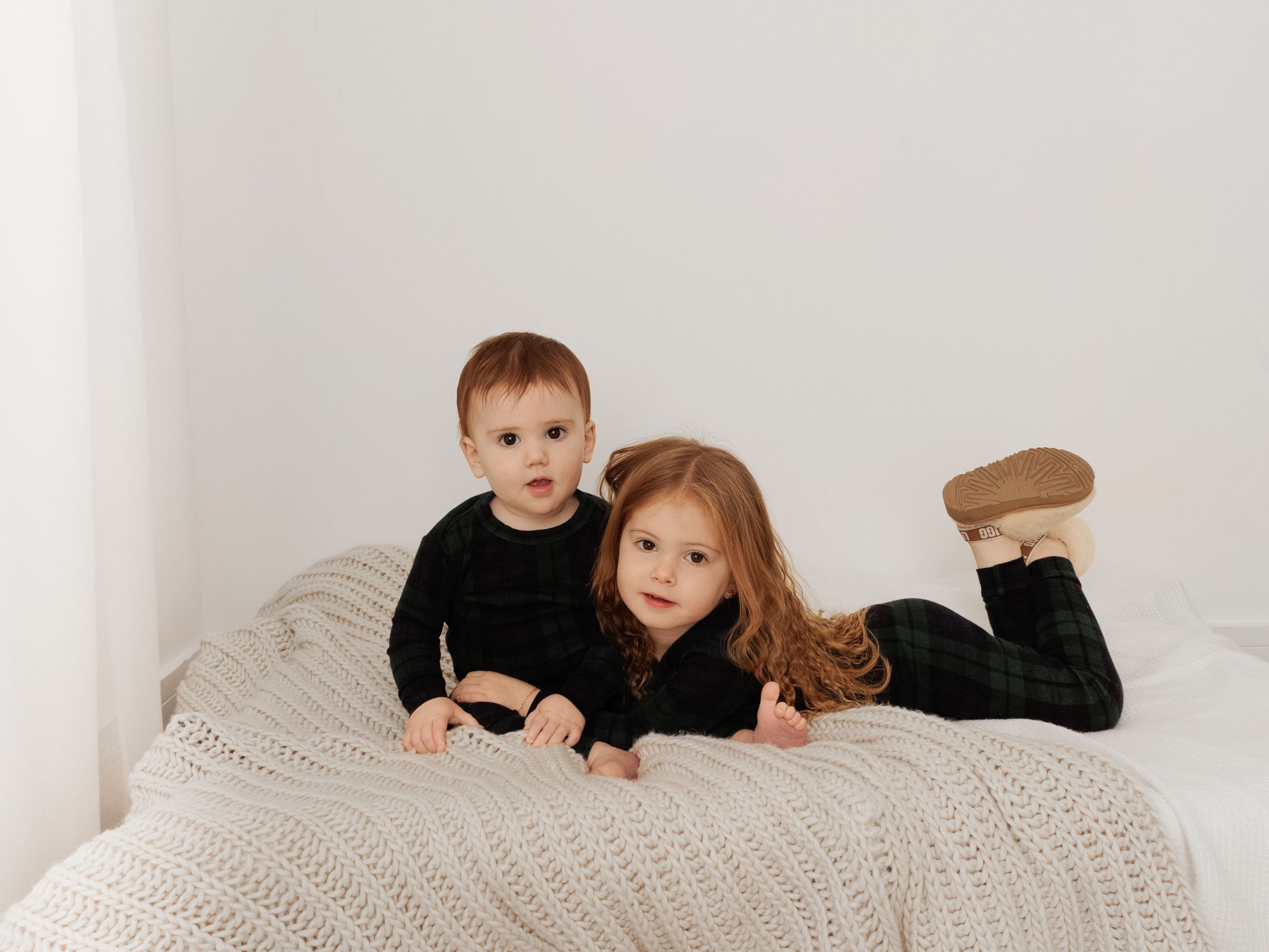 Two young children in dark outfits lying on a cream knit blanket against a white wall.