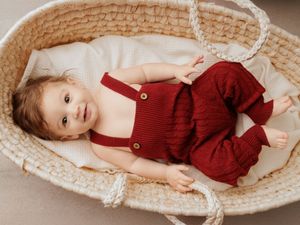 Baby in red knit overalls smiling while lying in a woven Moses basket with cream lining.