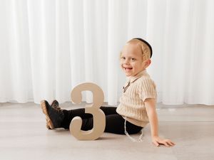 Smiling toddler boy sitting on floor beside a number 3, wearing a beige knit top and black pants for a birthday photo.