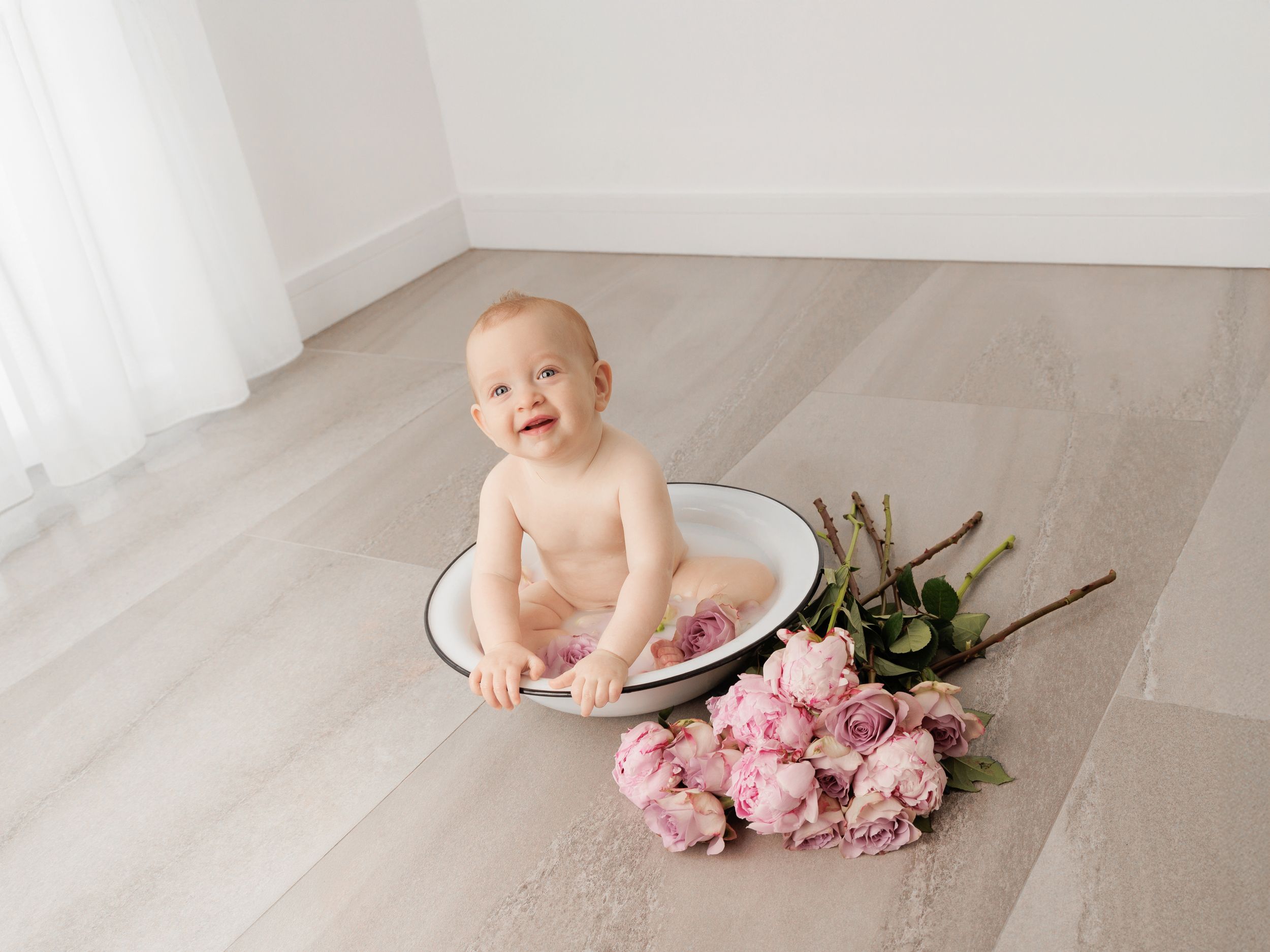 Baby sitting in a white bowl with pink roses on a light wood floor in a bright minimalist studio setting.