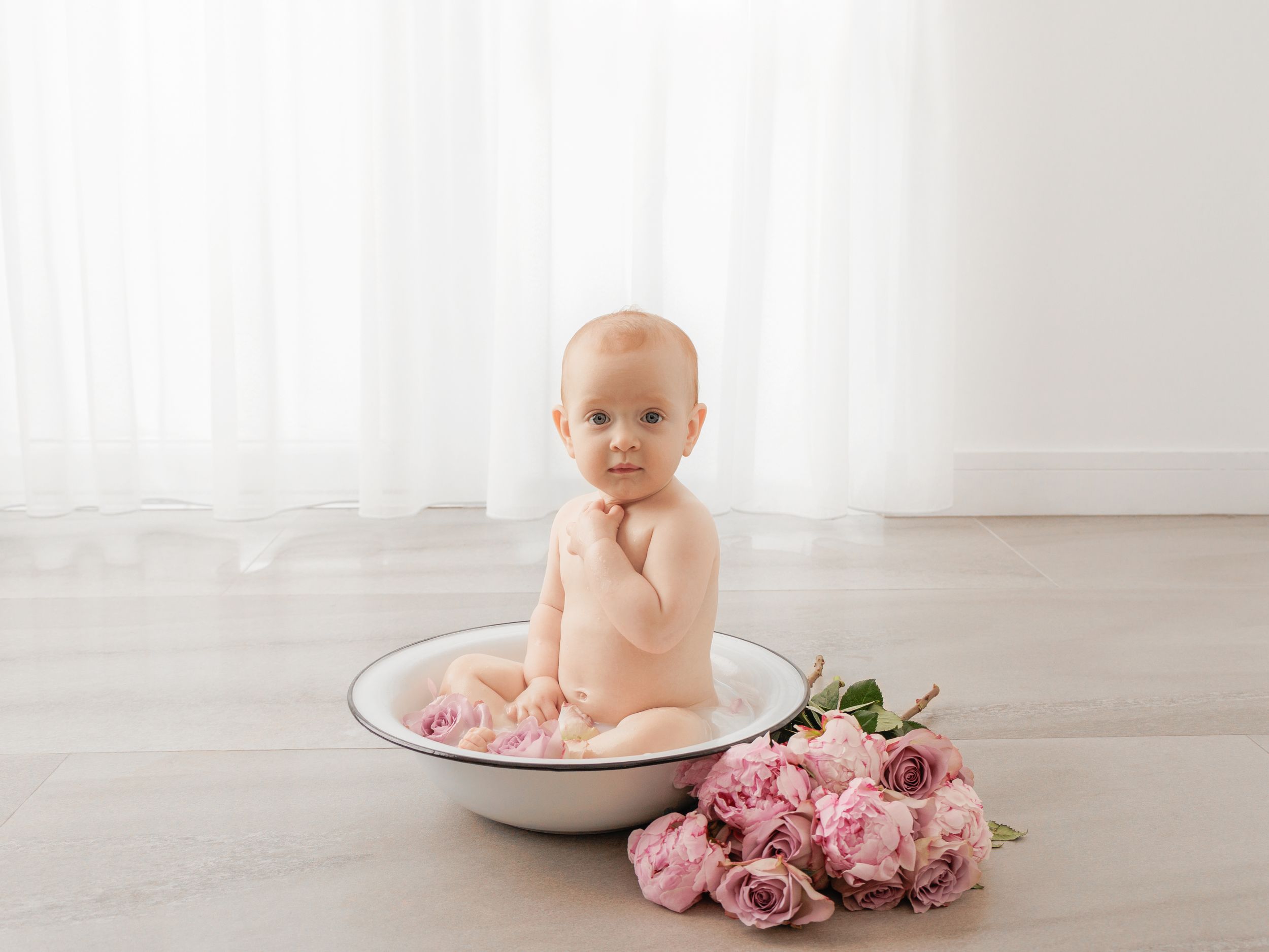 Baby sitting in a white enamel bowl surrounded by pink roses on a light wood floor with sheer curtains.