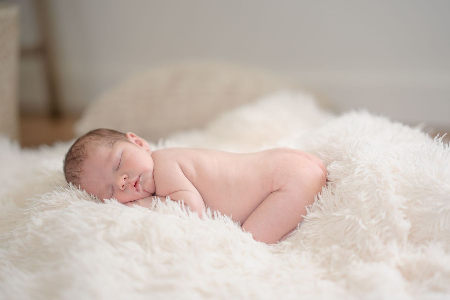 Peaceful newborn baby sleeping on a soft white fluffy blanket in a bright, airy room.