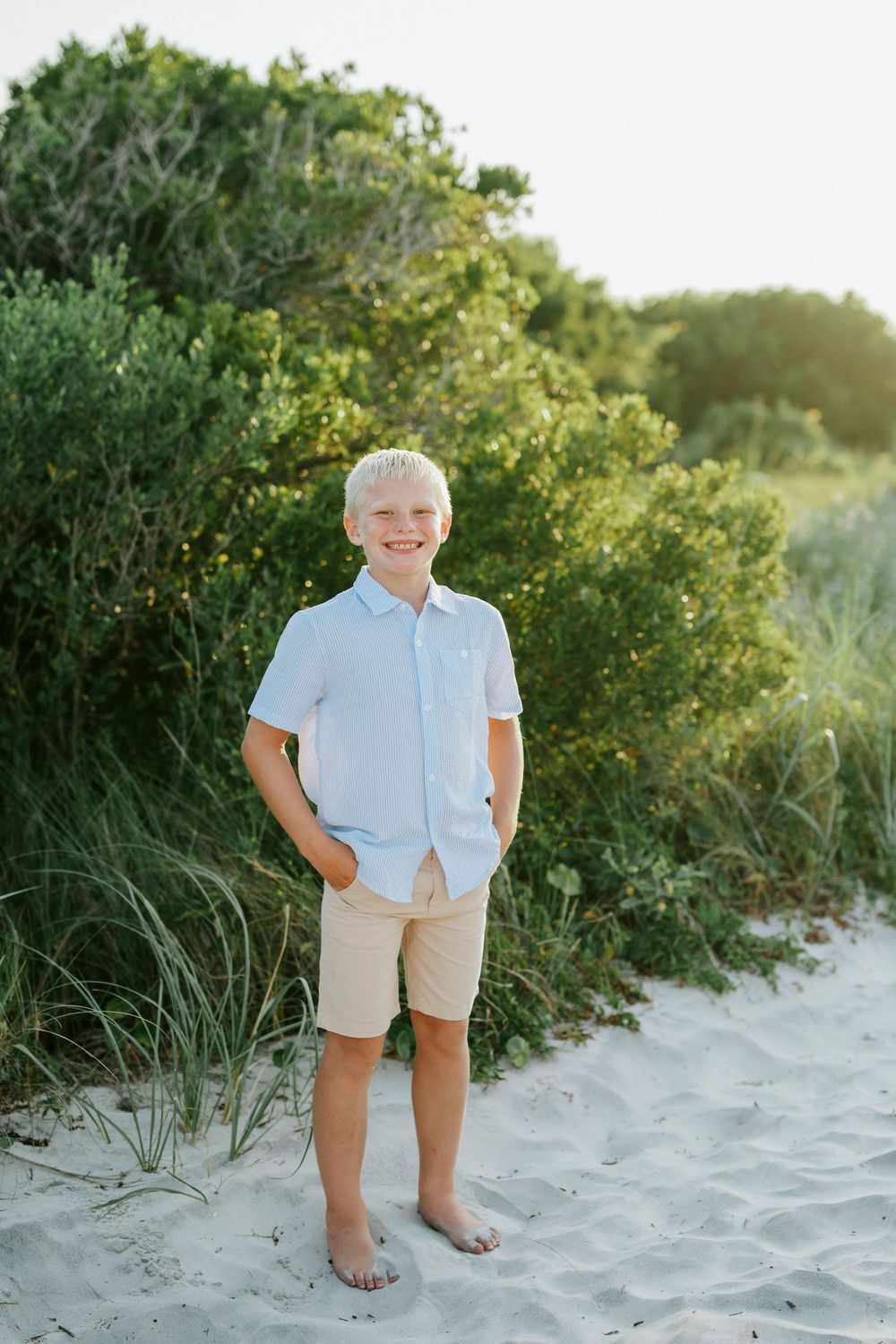 Smiling blond boy in light blue shirt and tan shorts standing barefoot on sandy beach with green bushes.