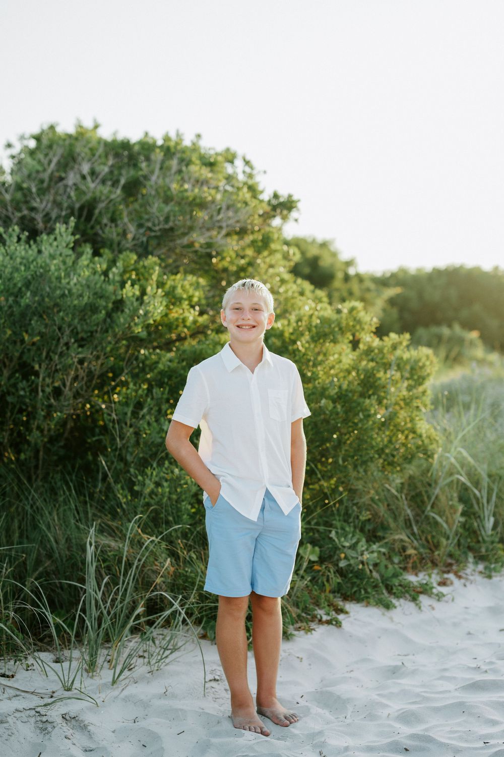 Smiling boy in white shirt and blue shorts standing barefoot on sandy beach path with green bushes behind him.