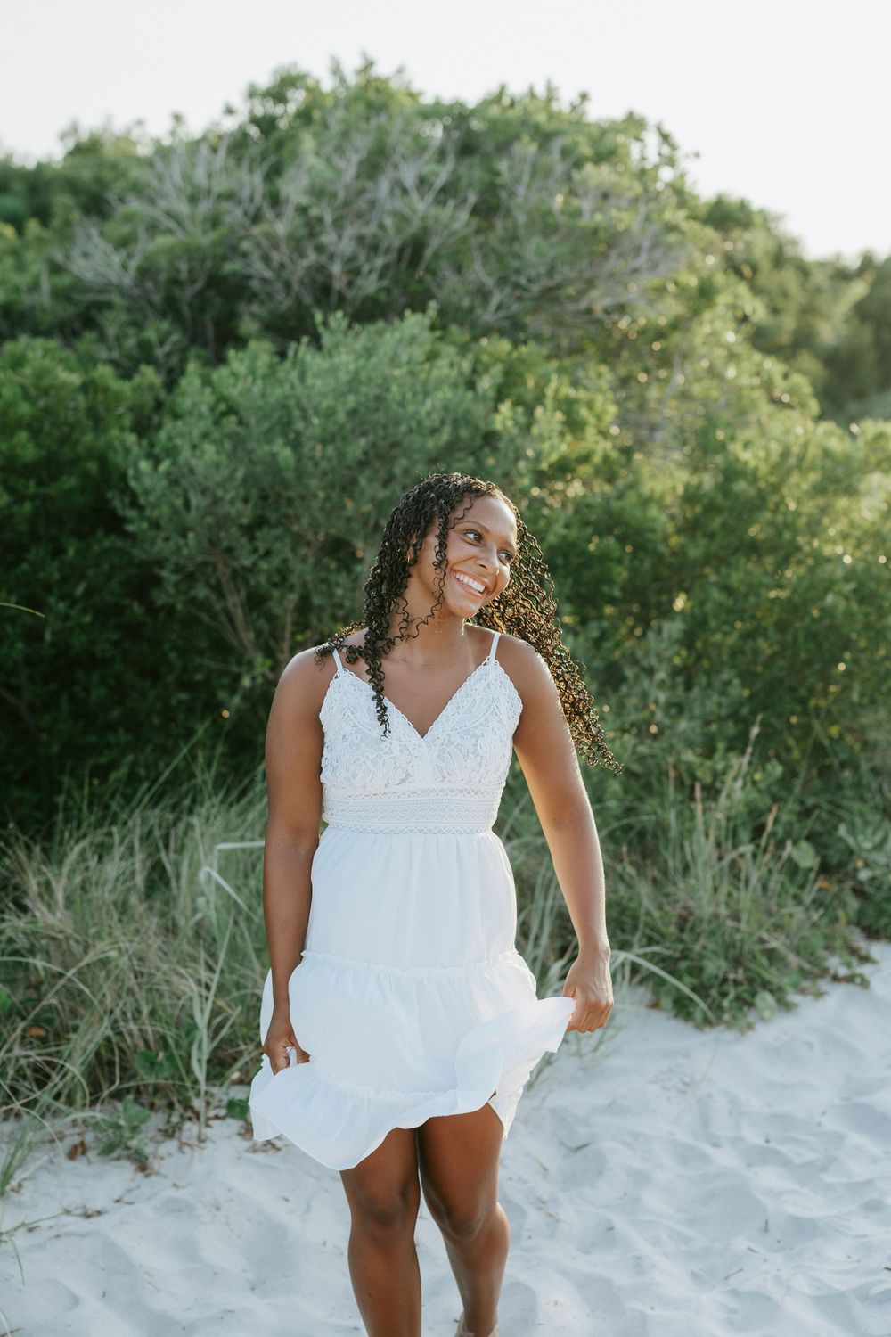 A smiling young Black woman in a white sundress stands on sandy beach dunes with lush green trees behind her.