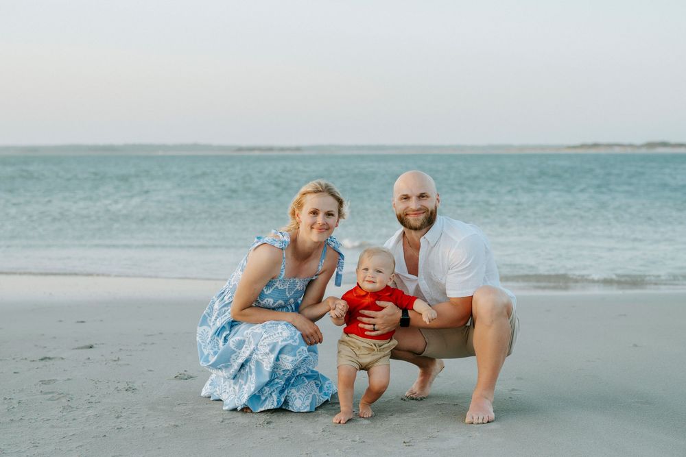 Family of three posing on sandy beach, parents smiling with baby in red shirt, ocean waves in background.