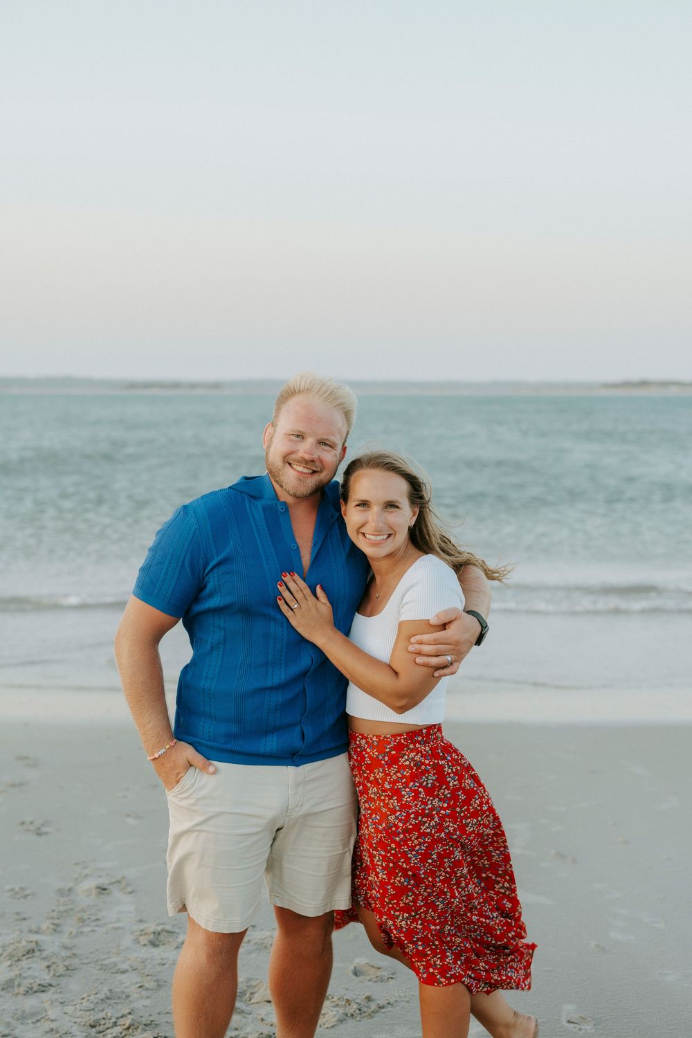 Happy couple embracing on a beach at sunset, man in blue shirt and woman in red floral skirt.