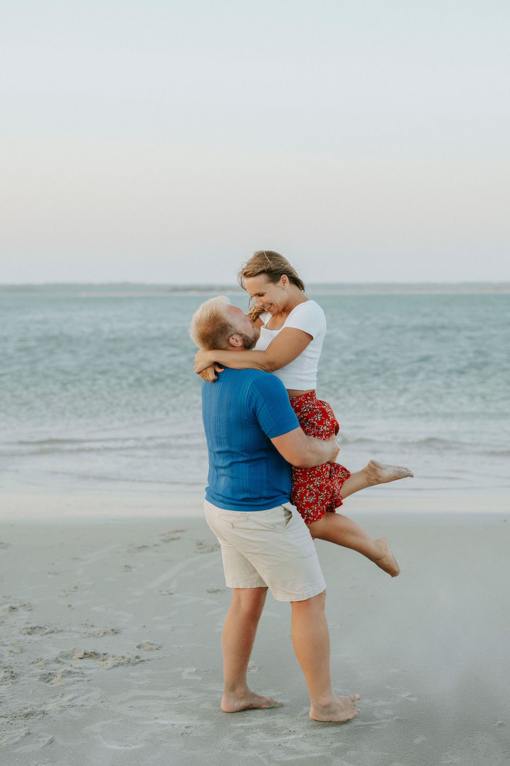 A man in a blue shirt lifts a woman in a red skirt on a sandy beach with ocean waves behind them.