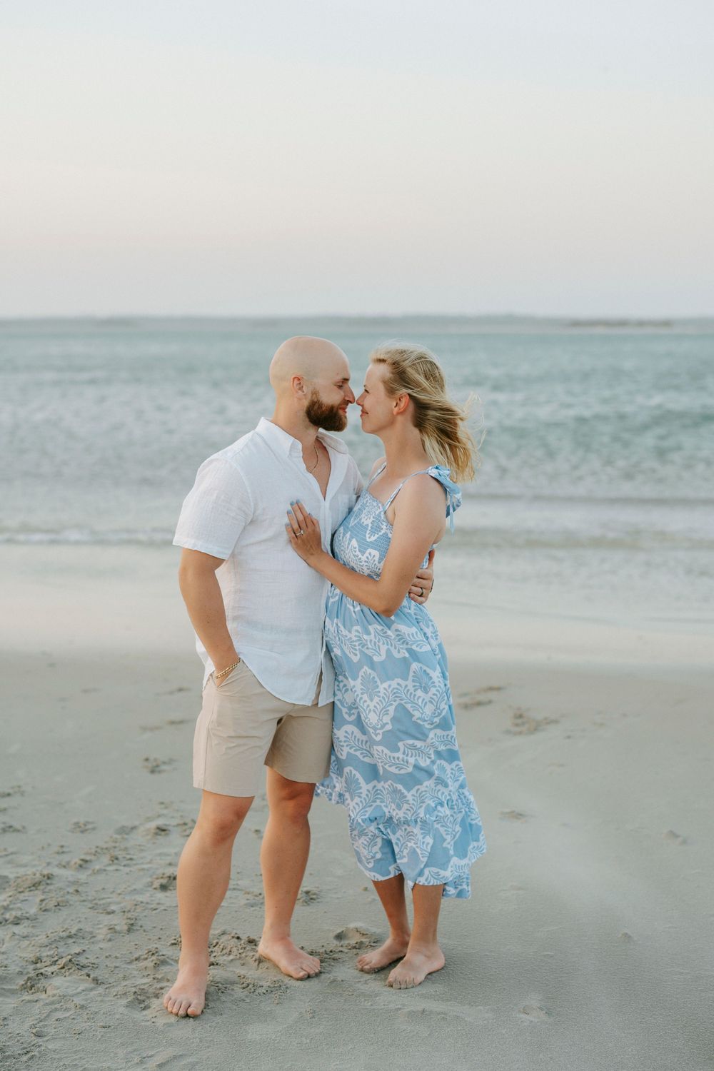 A couple shares a romantic moment on a sandy beach, the woman in a blue floral dress, the man in white shirt and khaki shorts.