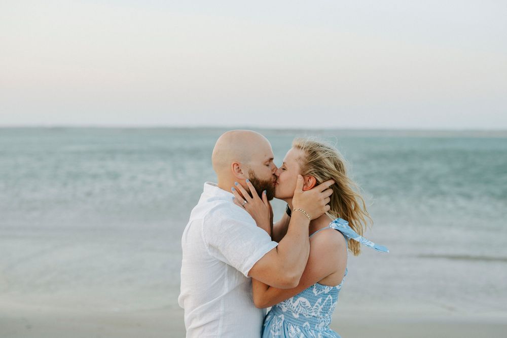 Couple sharing a romantic kiss on the beach with ocean waves in the background at sunset.
