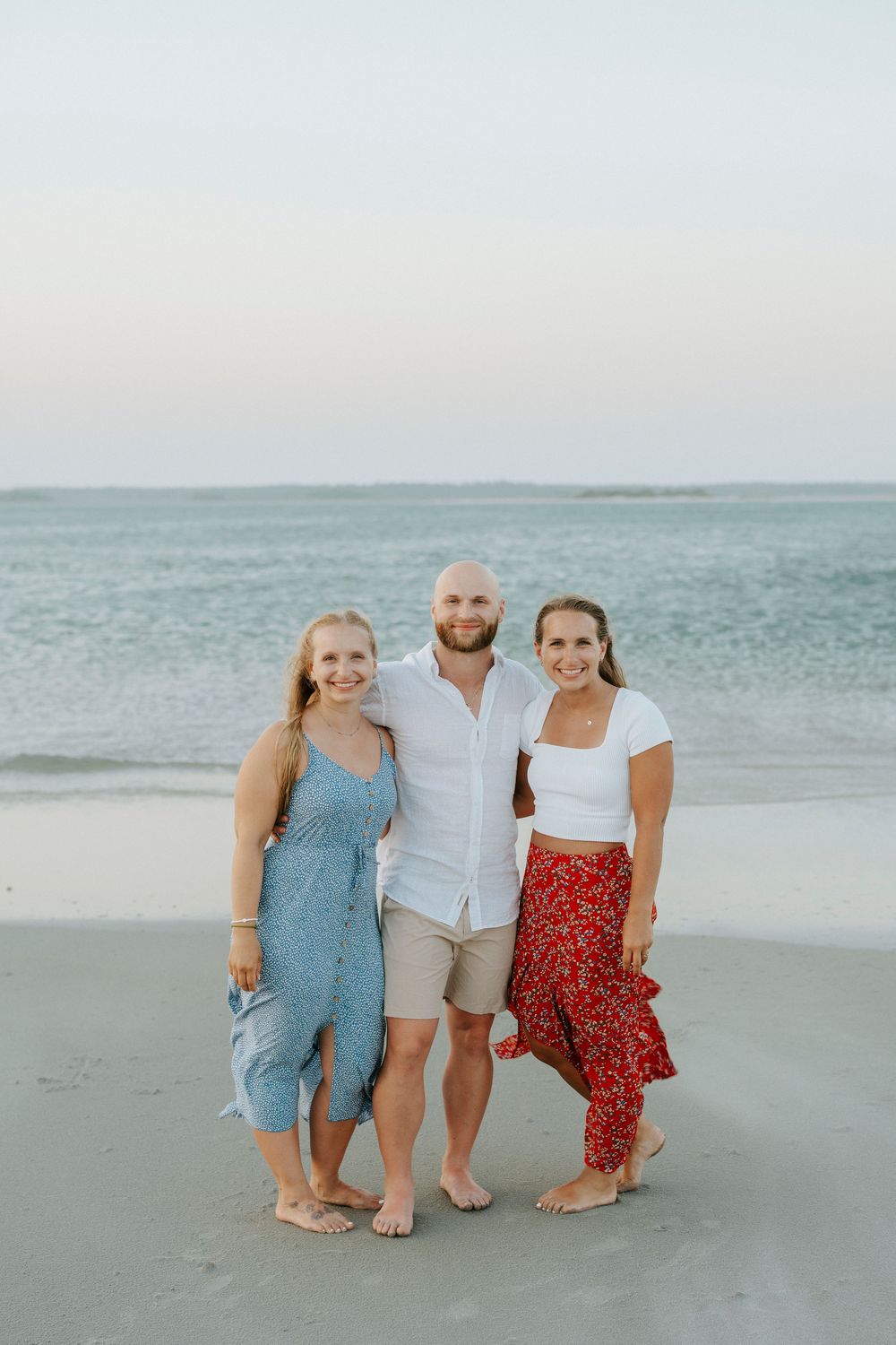 Three friends smiling together on a beach at sunset, man in white shirt with two women in summer dresses.