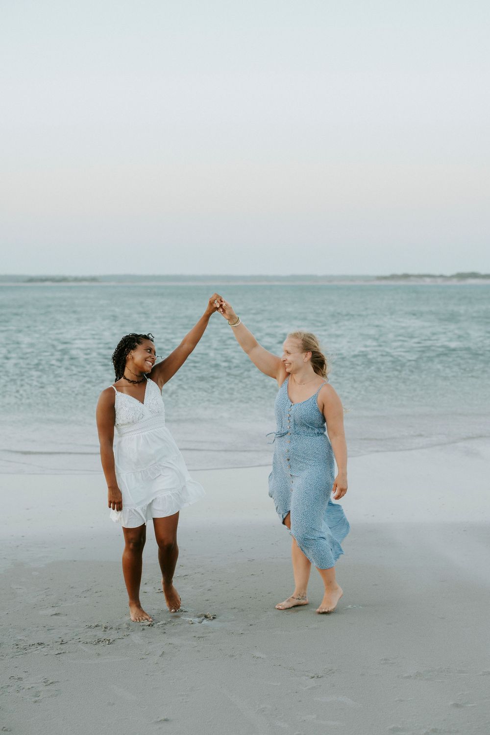 Two women in summer dresses dancing and holding hands on a sandy beach at dusk.