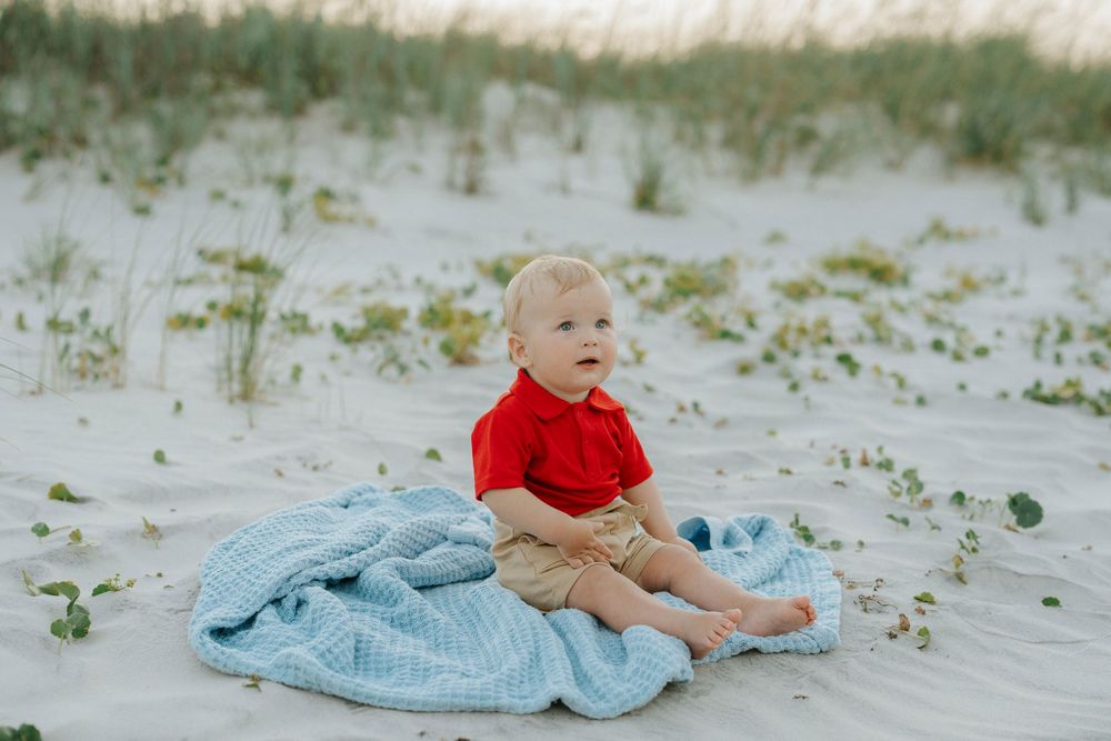 Baby in red shirt sits on blue blanket on white sand beach dunes.
