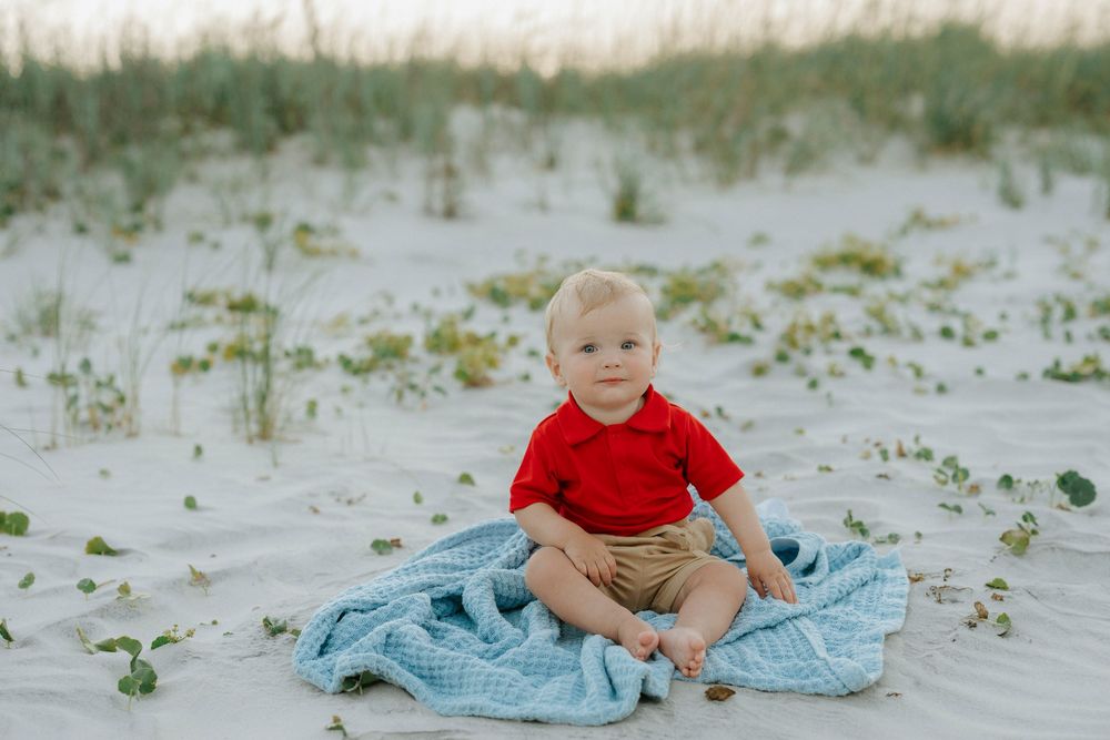 Toddler boy in red shirt sits on blue blanket on white sand beach dunes.