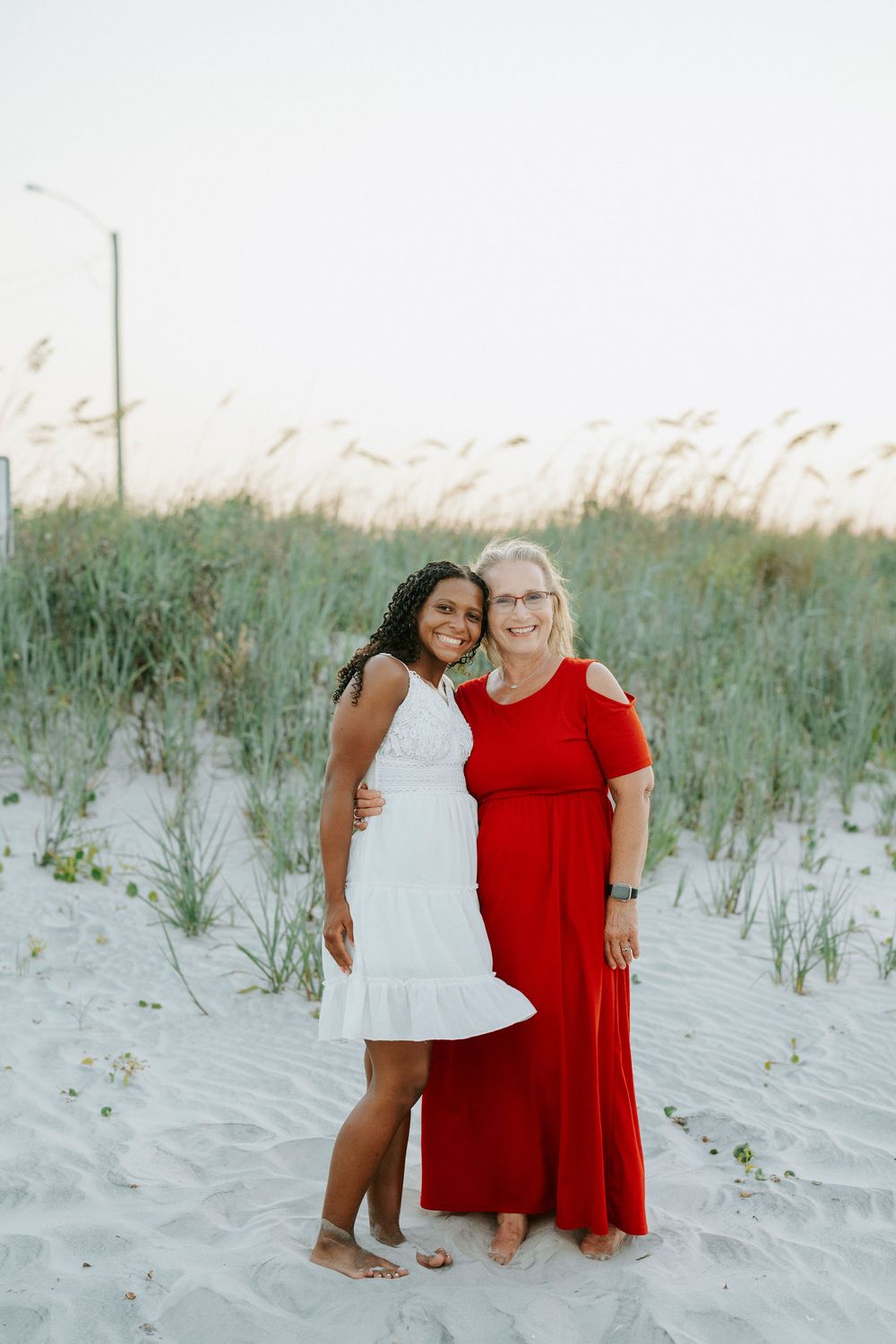 Two women in red and white dresses pose together on a sandy beach with sea oats in the background.
