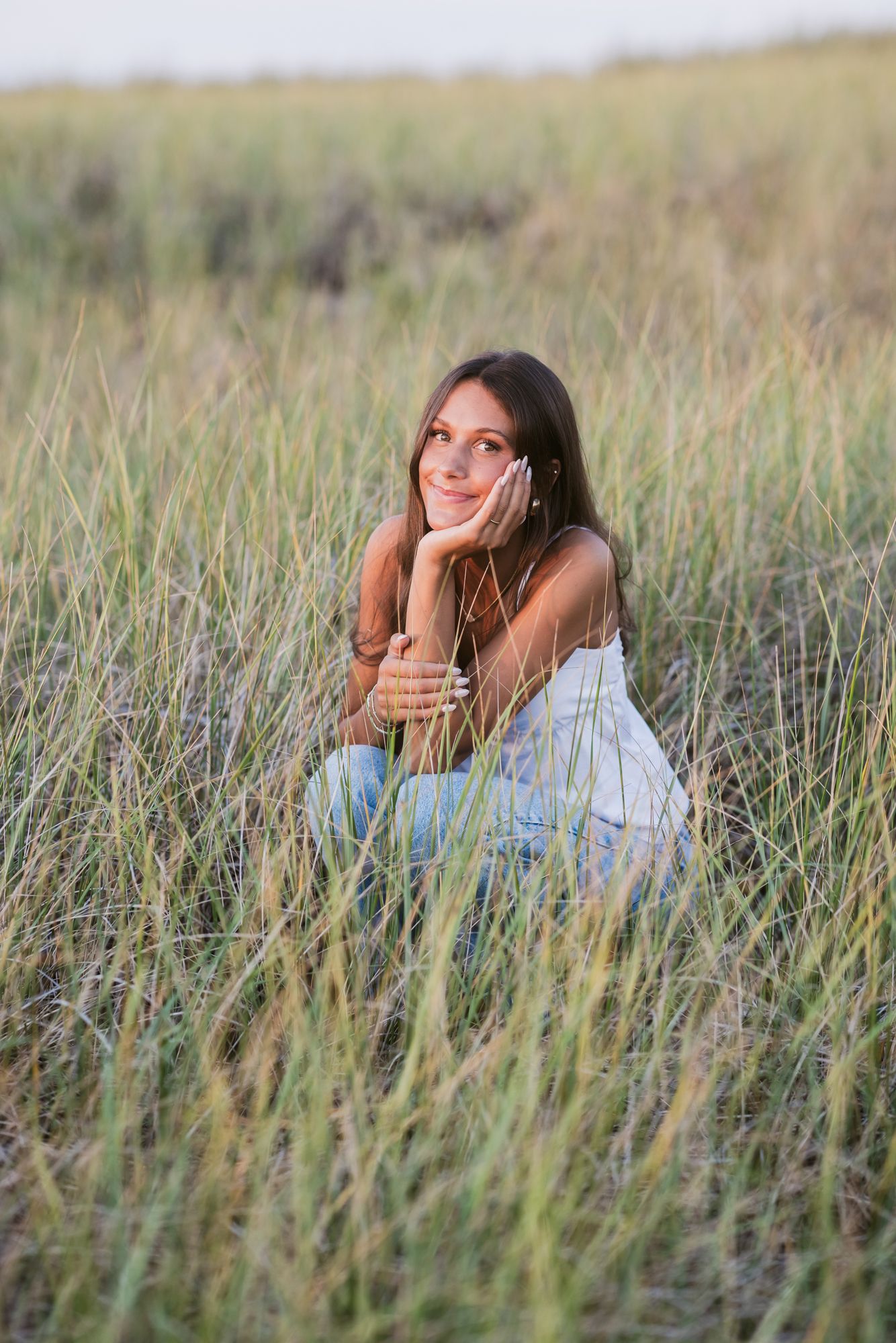 Teen wearing a white tank and light blue jeans squats in coastal grass for senior photos at Salisbury Beach. Seacoast senior photographer.