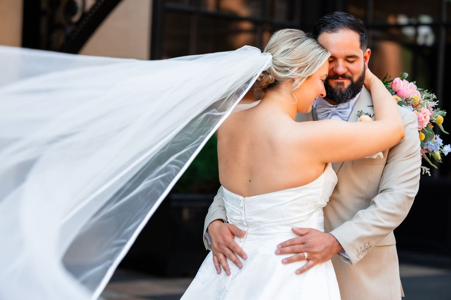 Joyful bride laughing with groom kissing her cheek, holding colorful bouquet under chandelier at elegant wedding venue.