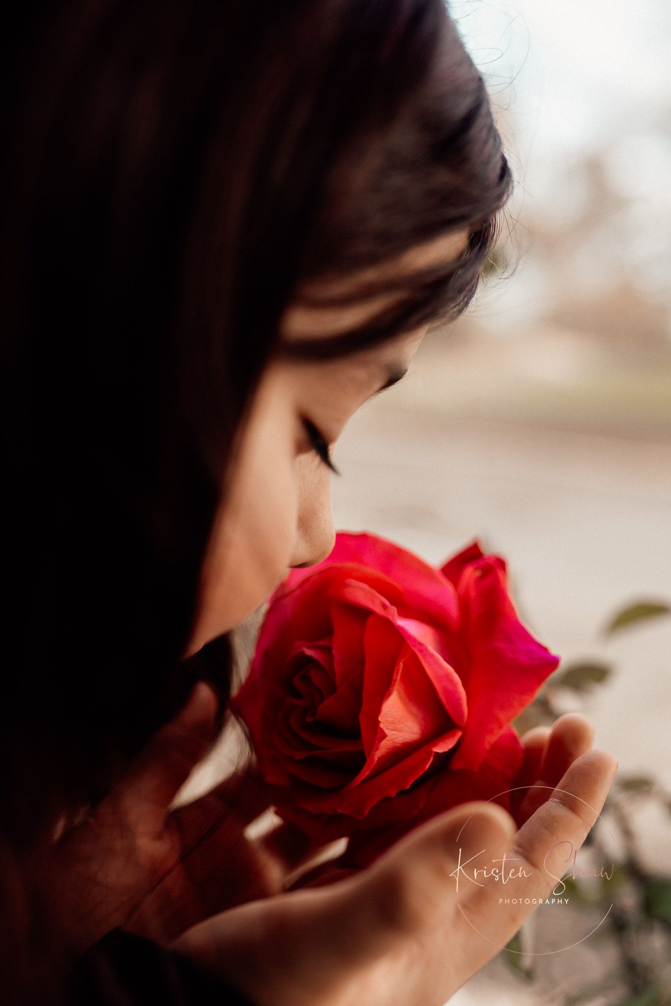 A young woman with dark hair smells a vibrant red rose held in her hands, captured in a moody portrait.