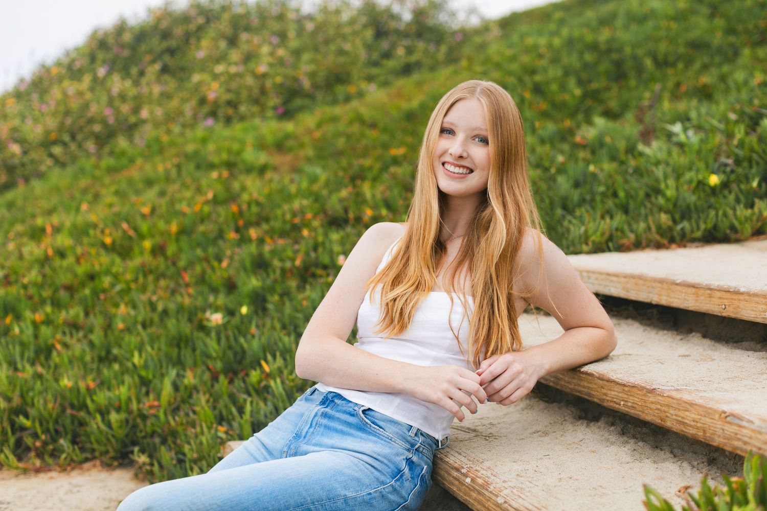 Young woman with long red hair smiling, sitting on wooden steps outdoors with green hillside background.