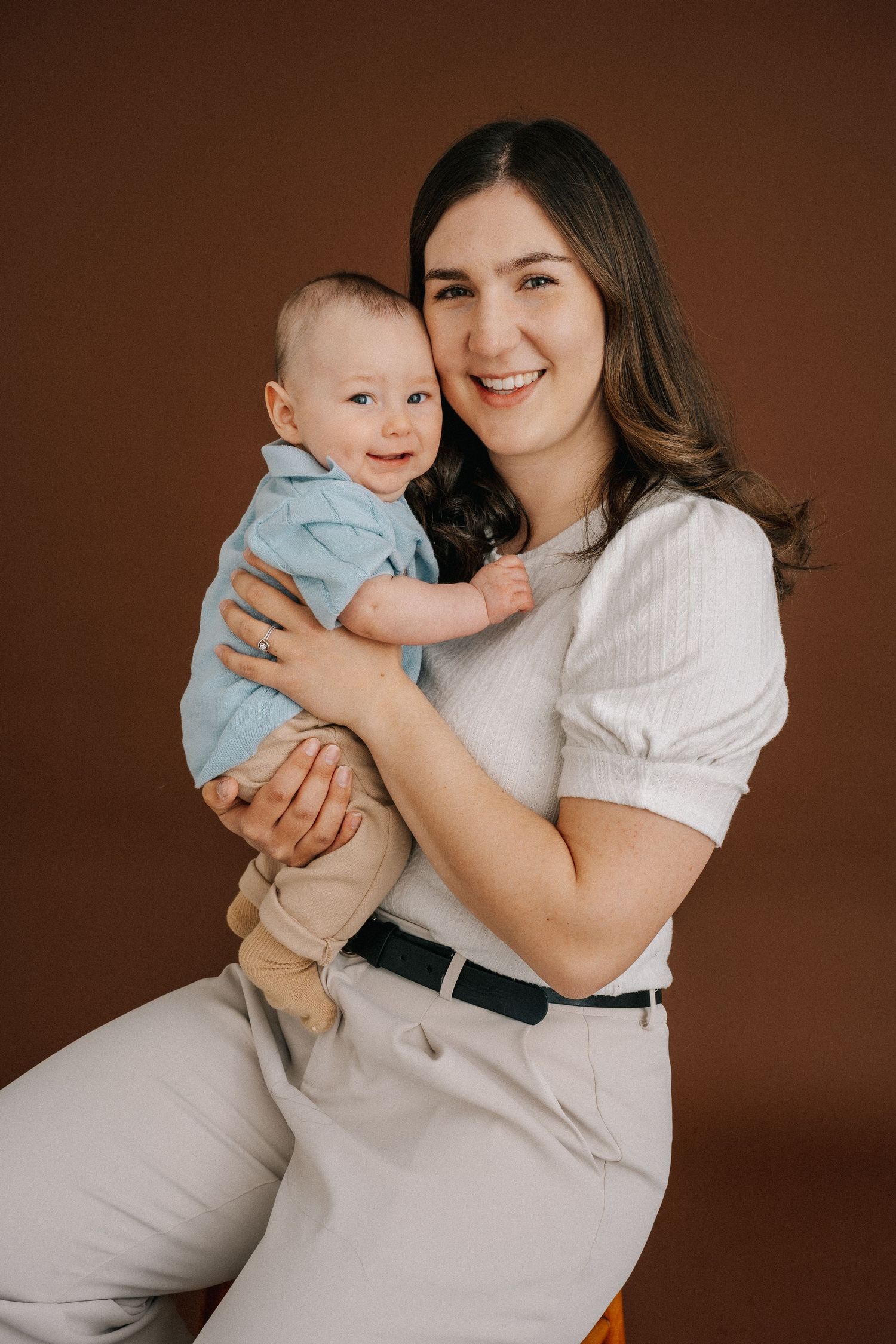 A smiling young mother holds her happy baby boy in a studio portrait against a warm brown background.