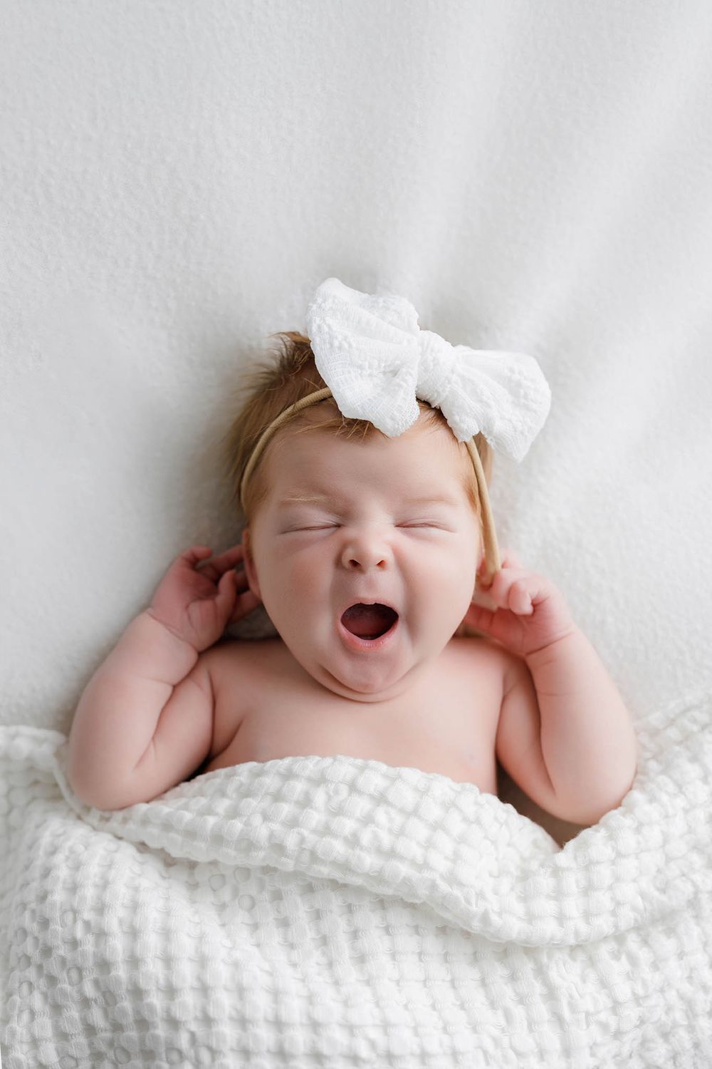 Adorable newborn baby girl yawning with white bow headband lying on soft white waffle blanket.