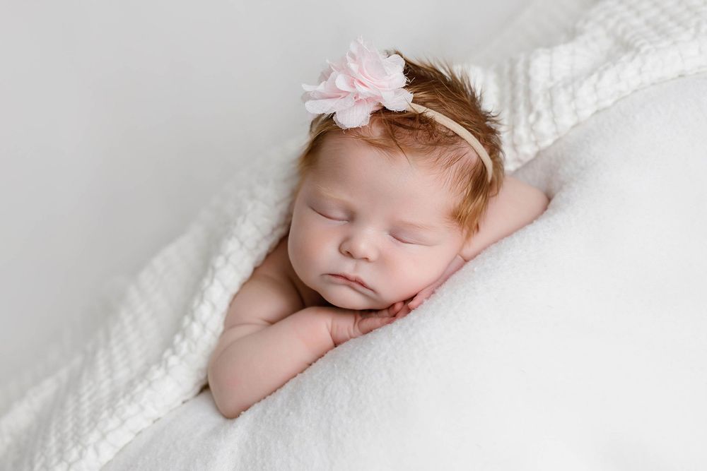 Peaceful newborn baby girl sleeping on white blanket wearing a delicate pink flower headband.