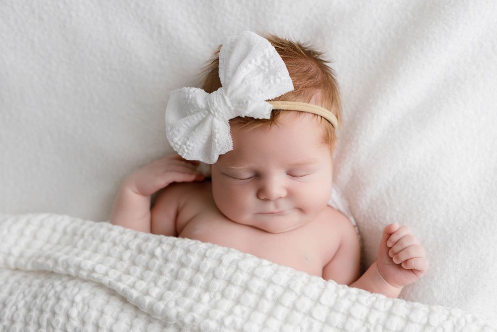 Peaceful newborn baby girl sleeping with white lace bow headband wrapped in soft white knit blanket.