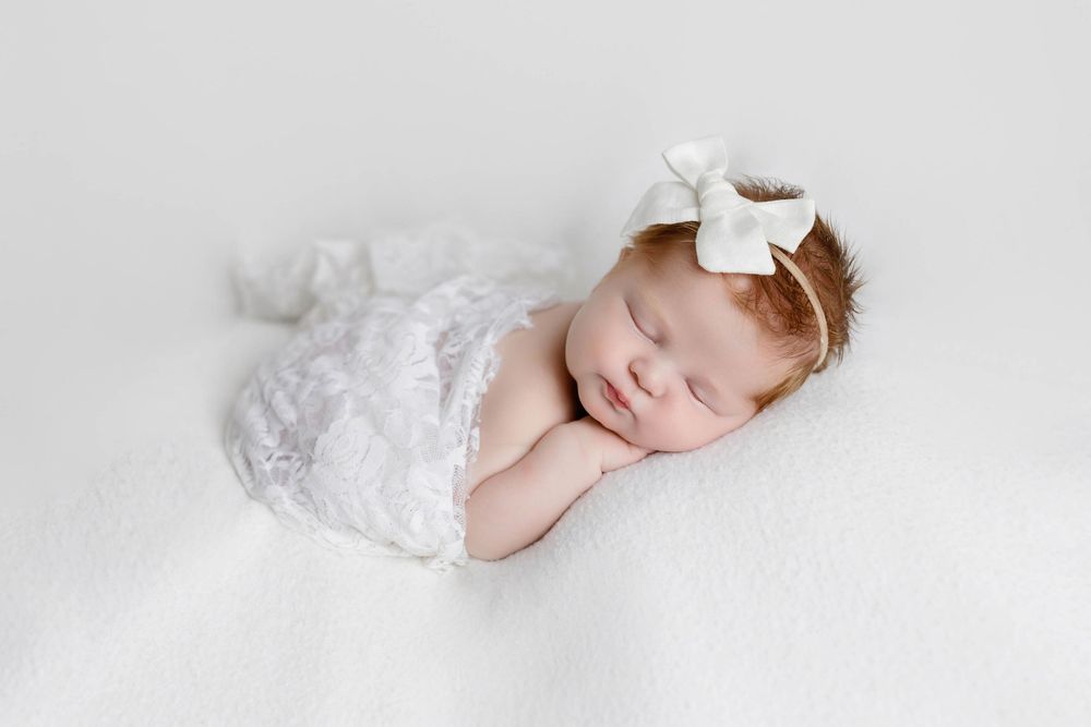 Sleeping newborn baby girl in white lace romper and bow headband on soft white blanket.
