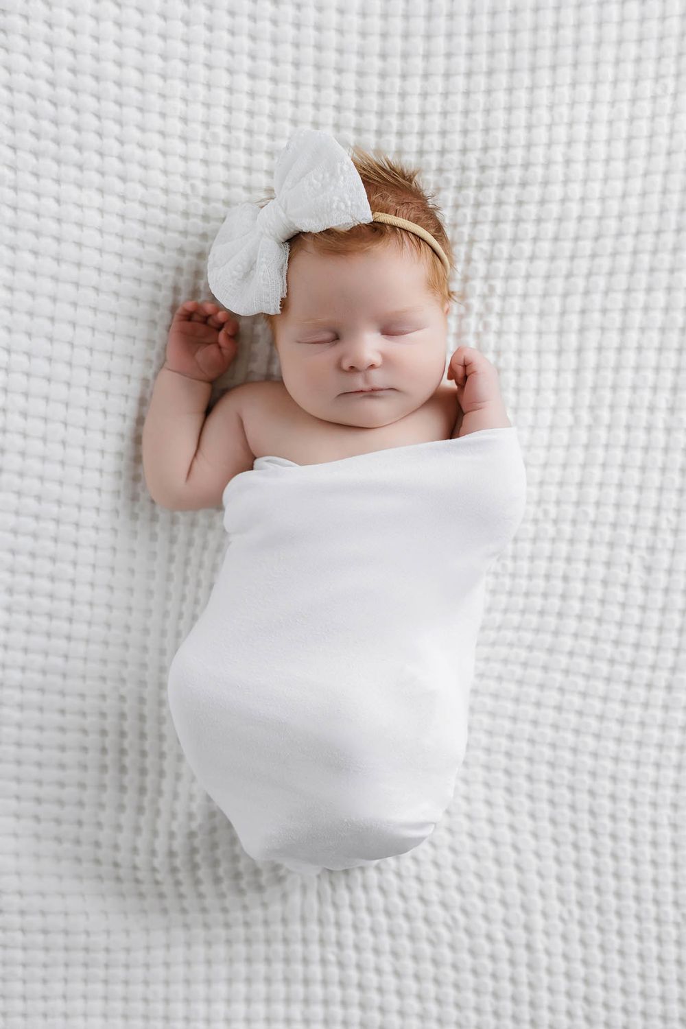Peaceful newborn baby girl swaddled in white blanket with bow headband sleeping on white textured background.