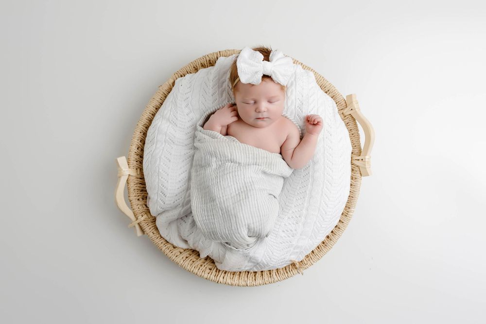 Newborn baby girl wrapped in white blanket inside wicker basket with bow headband.