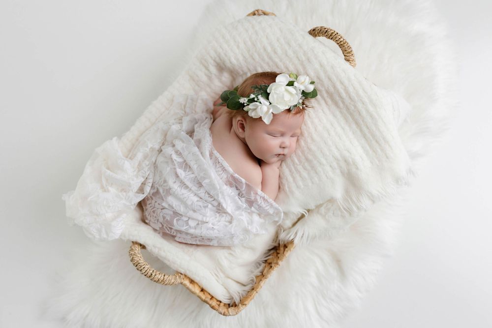 Newborn baby girl sleeping in wicker basket wrapped in white lace with floral crown on white fur backdrop.
