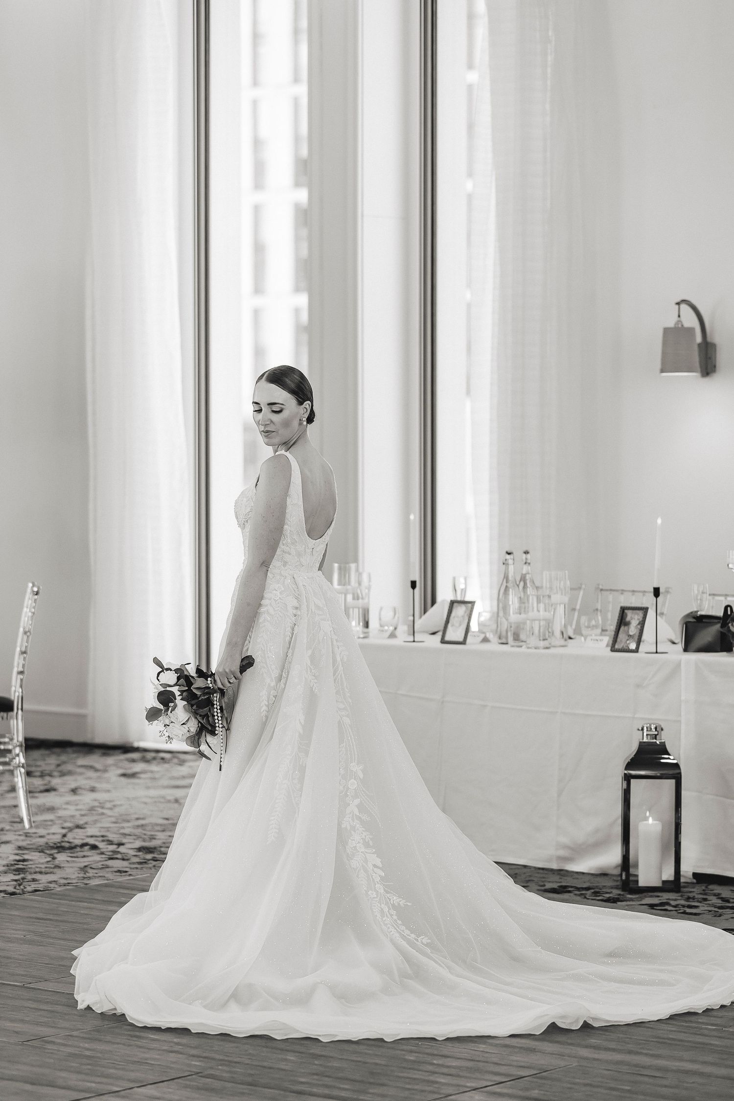 Black and white photo of a bride in an elegant gown with a long train, holding a bouquet at a wedding reception.