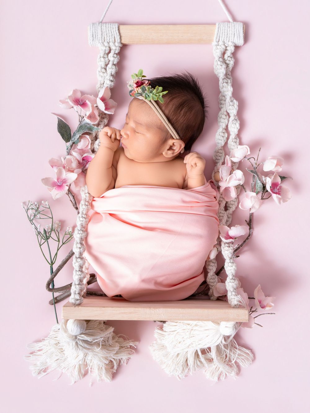 Newborn baby sleeping peacefully wrapped in pink on a macrame swing with floral decorations.