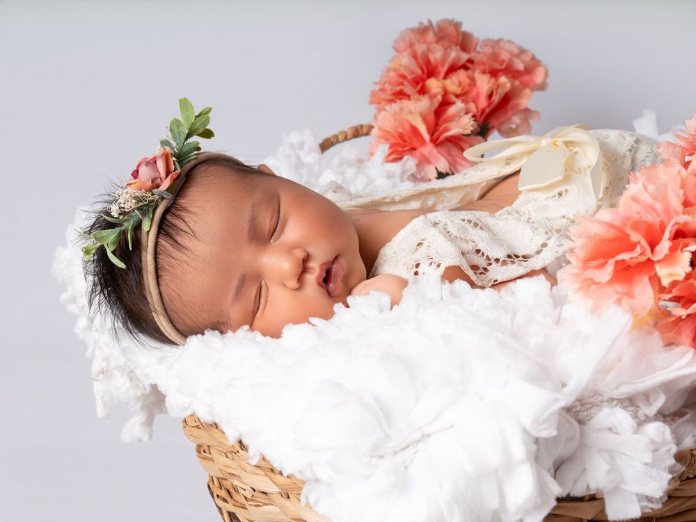 Sleeping newborn baby in a wicker basket with white ruffles, pink flowers, and a floral headband.