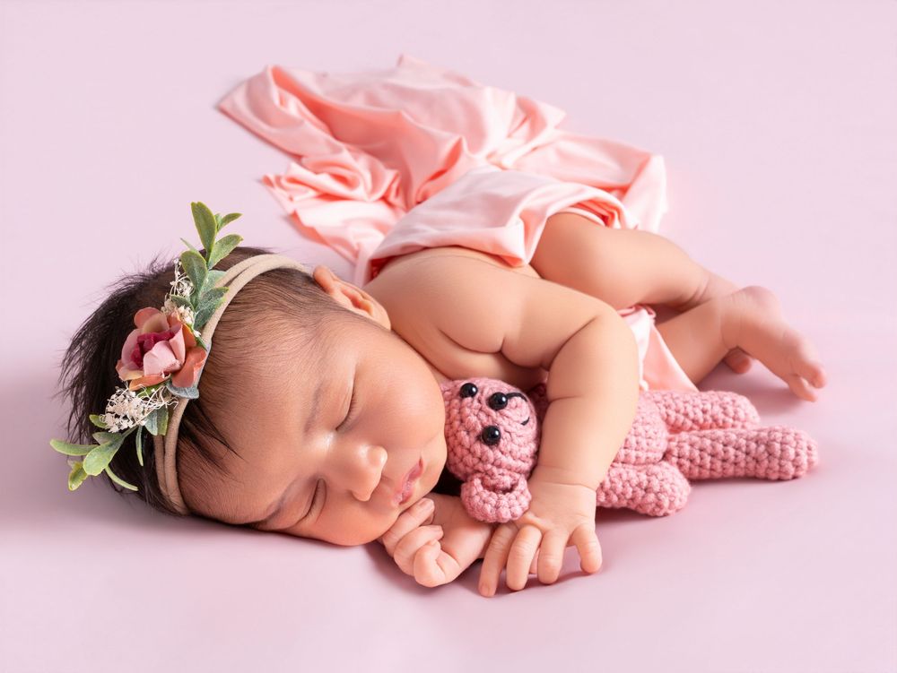 Sleeping newborn baby girl in pink dress and floral headband cuddling a pink crochet teddy bear on pink background.