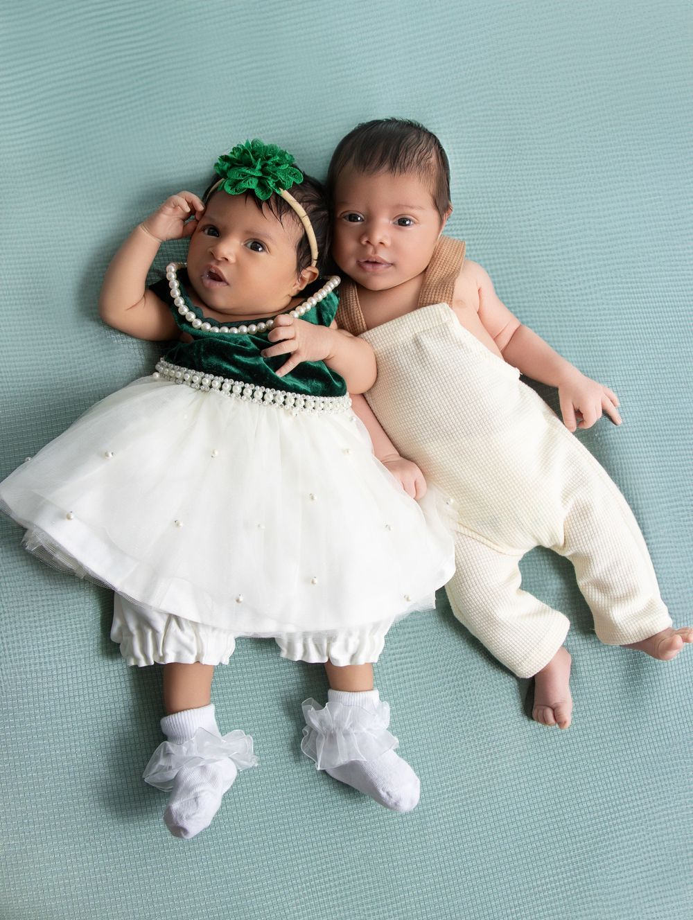 Two adorable babies in white outfits lying together, one wearing a green floral headband and pearl-accented dress.