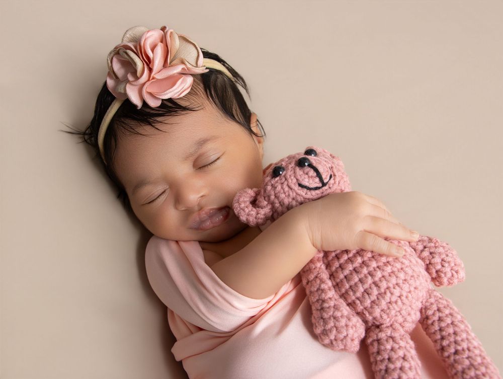 Sleeping newborn baby girl in pink wrap holding a pink crocheted teddy bear with a floral headband.