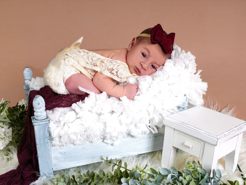 Newborn baby in lace outfit and red bow headband posed on miniature white bed with fluffy white blanket.