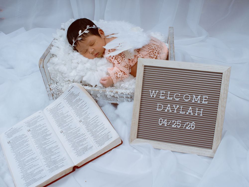 Newborn baby girl in pink lace outfit sleeping in basket beside open Bible and Welcome Daylah 04/25/26 letter board.