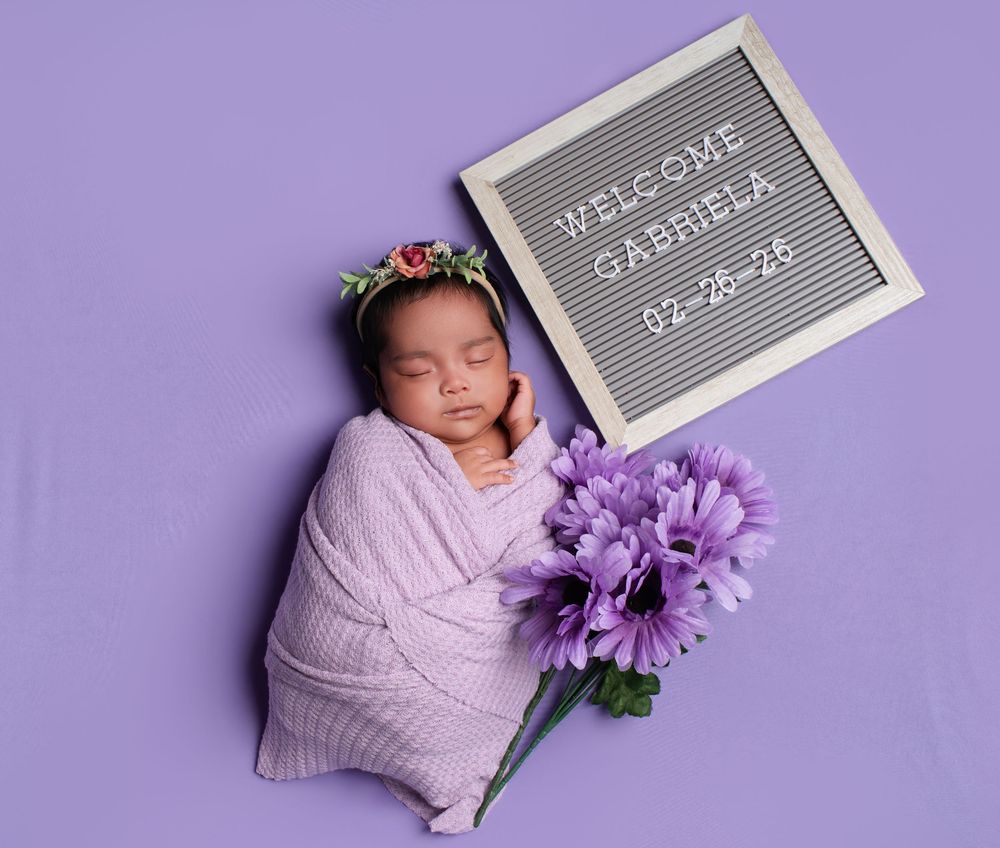 Newborn baby girl Gabriela swaddled in purple wrap with floral headband beside welcome sign and purple flowers.