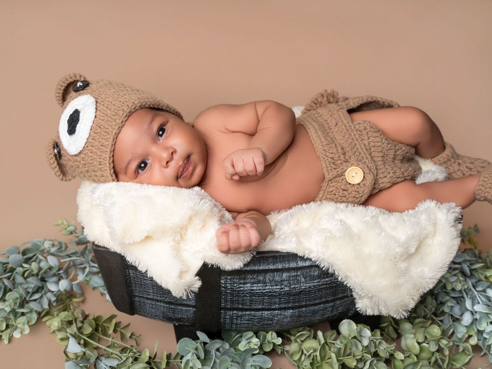 Smiling newborn baby in knit bear hat and outfit resting in a small bucket with greenery.